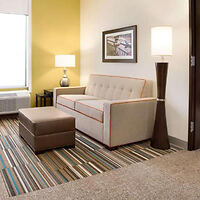 A modern living room with a beige sofa, a brown ottoman, a side table with a lamp, a standing lamp, and a framed picture on the wall. The floor is carpeted with striped patterns.