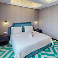 A modern bedroom with a large bed featuring white linens, a teal headboard, two bedside tables with lamps, and a green and white chevron-patterned rug. Folded towels are placed on the bed.