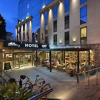 A modern hotel entrance at dusk, featuring a glass façade, outdoor dining area, bike rack, and illuminated "Hotel Loft" signage. Several people are seated at tables, enjoying their meals.