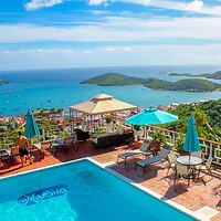 A hillside infinity pool with lounge chairs and umbrellas overlooks a scenic ocean view with distant islands and boats.