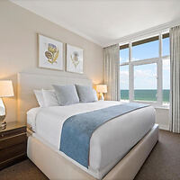 A bedroom with a large bed featuring white linens and a blue throw, flanked by two nightstands with lamps. Large window with sea view, two framed artworks above the bed. Neutral-toned decor.