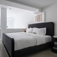 A modern, minimalist bedroom with a large black-framed bed, white linens, a bedside table, a vase of flowers, and a framed abstract art piece on the wall.