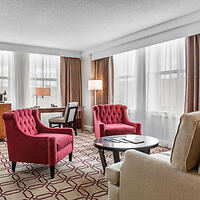 A spacious hotel room features two red armchairs, a beige chair, a coffee table, desk, and large windows with sheer curtains, letting in natural light.