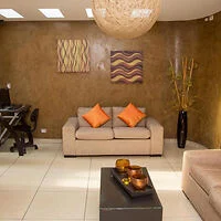 A modern living room with a beige sofa, orange cushions, abstract wall art, a black coffee table, and a desk with a computer on the side. A large, spherical light fixture hangs from the ceiling.