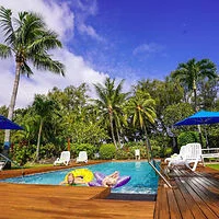 Person lounging on a float in a pool surrounded by tropical trees and blue umbrellas, under a clear blue sky.