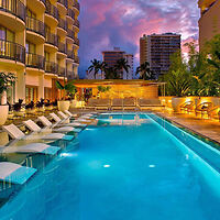 Poolside view at a luxury hotel with lounge chairs, illuminated pool, and palm trees, set against a backdrop of a vibrant sunset sky and cityscape.