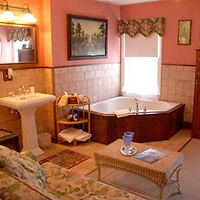 A cozy bathroom with a pedestal sink, freestanding bathtub, and a small shelving unit. There is a sofa and a coffee table with a blue vase on it. Walls have artwork and a window with a patterned valance.