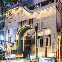 A modern building with a unique facade featuring large arched windows and illuminated architectural details. It is situated on a bustling urban street lined with trees and colorful signage.