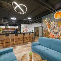 A modern lounge area with blue sofas, a wooden coffee table, and barstools at a counter. There is abstract wall art, decorative lighting, and a mix of contemporary and industrial design elements.