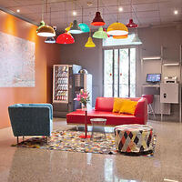 A colorful lounge with a red sofa, blue chair, multi-colored ottoman, vending machine, and hanging pendant lights. There is a vibrant rug, and a wall-mounted touchscreen is visible in the background.