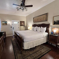 A neatly arranged bedroom with a wooden bed, white bedding, and two bedside tables with lamps. There is a ceiling fan above and a carpet under the bed, with light coming from a window on the left.