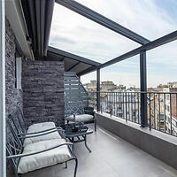 Covered balcony with metal railing, two cushioned chairs, a small table, and a view of city rooftops under a partly cloudy sky.