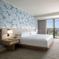 Modern hotel room with a king-size bed, blue patterned wallpaper, wooden nightstand with lamps, and floor-to-ceiling window offering a view of the outdoors.