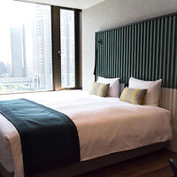 A hotel room with a double bed, green headboard, white linens, and green pillows. Large window with city skyline view.