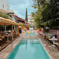 Outdoor space with a narrow swimming pool, surrounded by seating areas with umbrellas and colorful decor. Tables and chairs are set up on the left side, and a string of lights hangs above.