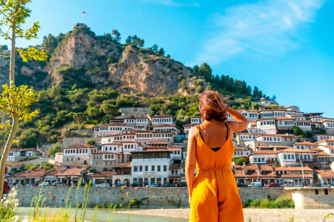 A person in an orange dress stands looking at a hillside town with white houses and a rocky hill in the background under a clear blue sky.