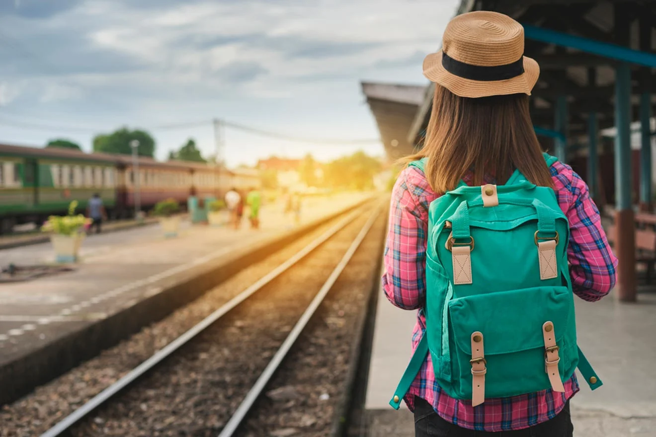 A person wearing a hat and green backpack stands on a railway platform, looking at the train tracks as the sun sets in the background.