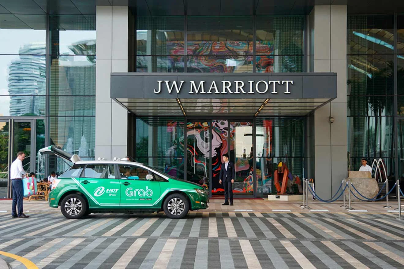 A green Grab taxi is parked outside the entrance of the JW Marriott hotel, with hotel staff assisting a guest at the entrance.