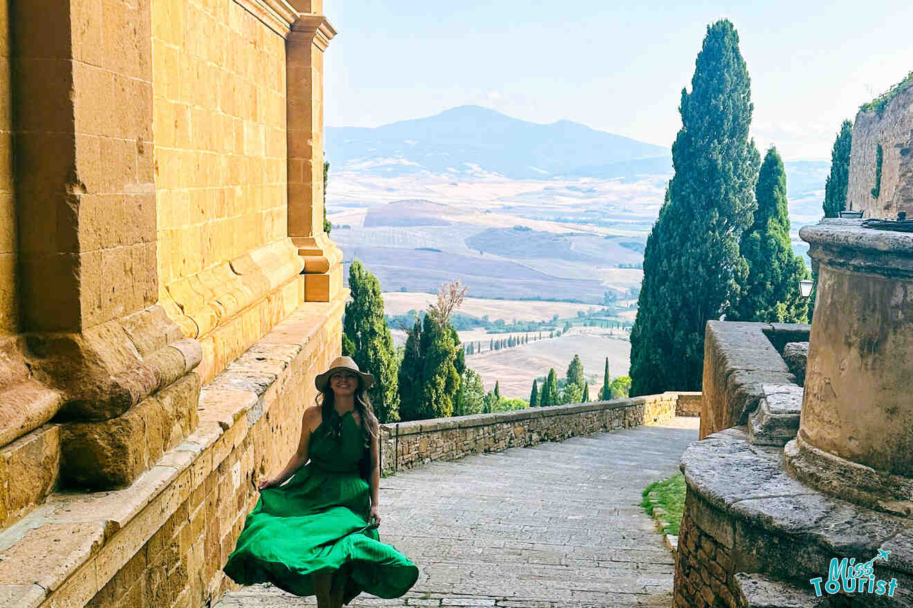 The writer of the post in a green dress walks down an ancient stone pathway in Pienza, with a backdrop of Tuscan hills and cypress trees in the distance.