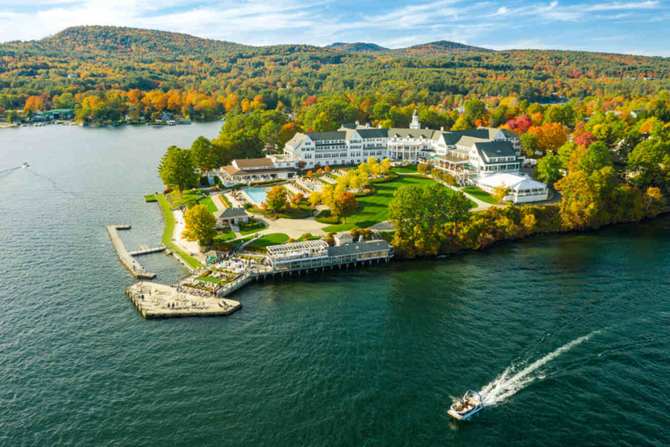 An aerial view of a lakeside resort surrounded by vibrant autumn foliage, with a boat on the lake and a forested hillside in the background.