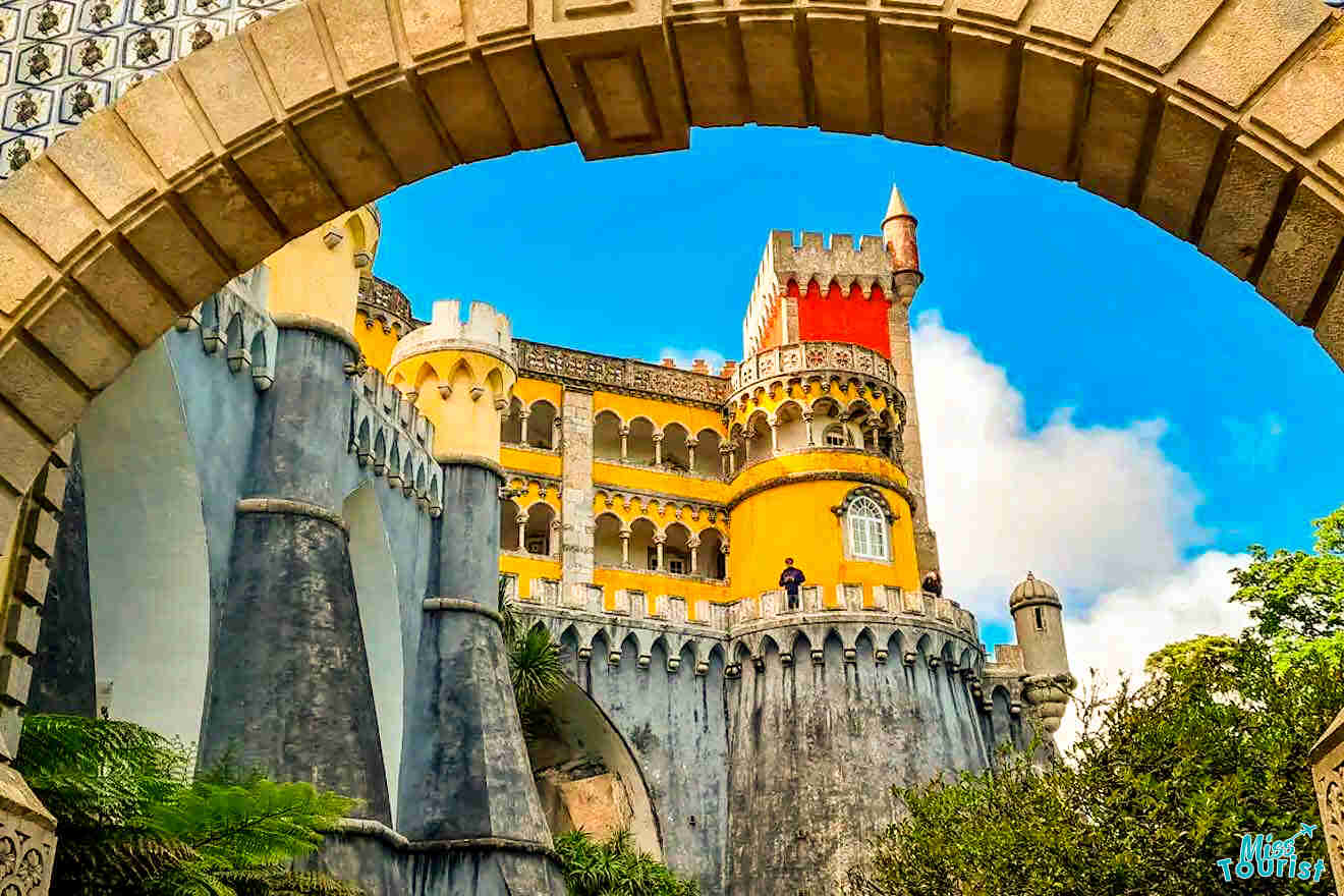 A colorful castle in Portugal features yellow, red, and gray towers framed by an arch beneath a vibrant blue sky with clouds.