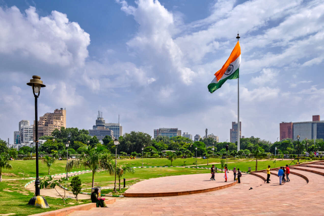 A large Indian flag flying in a park in Connaught Place, New Delhi, with city buildings in the background.