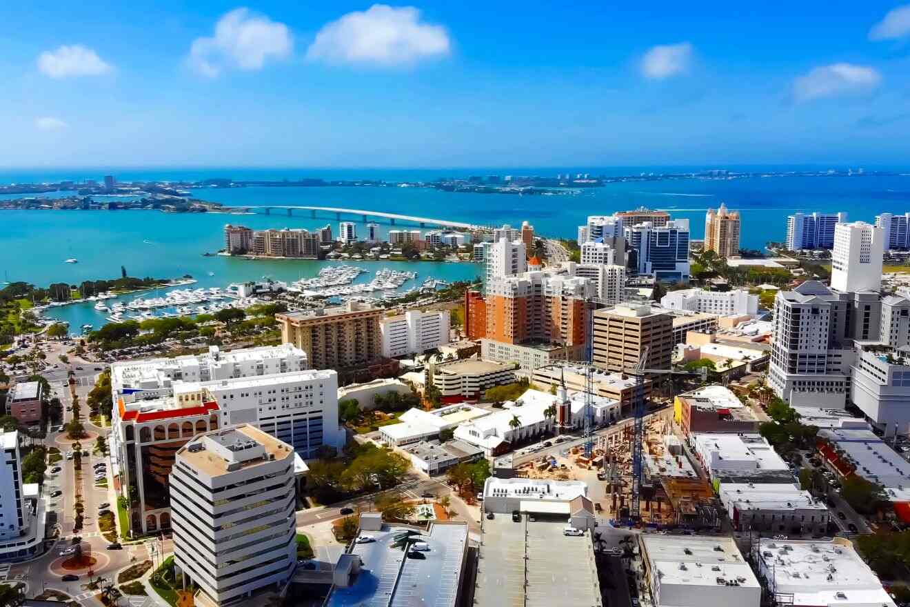 A panoramic view of downtown Sarasota, showing high-rise buildings, a marina filled with boats, and a bridge extending over the blue waters to a distant shoreline under a bright blue sky.