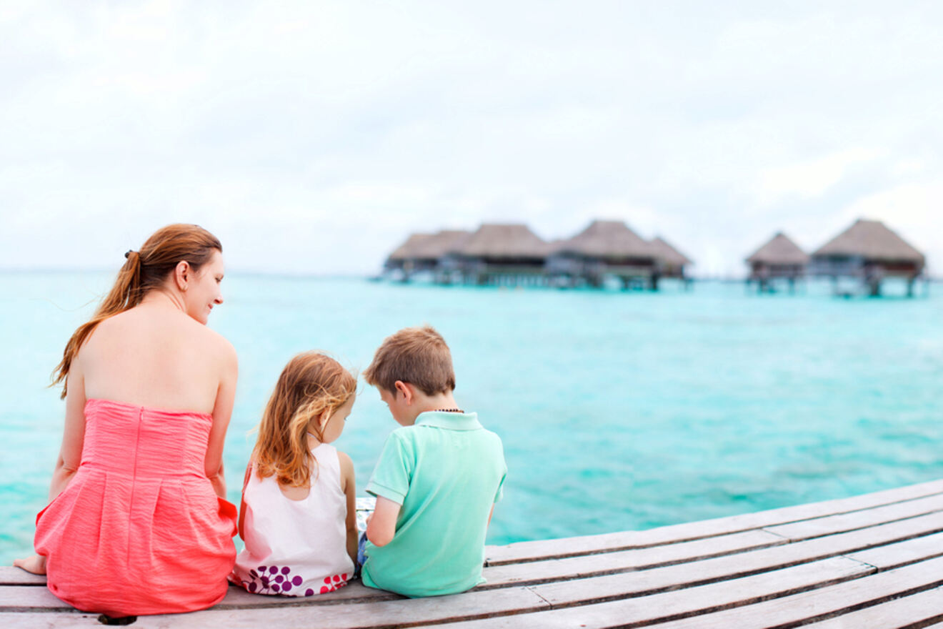 A woman and two children sit on a wooden deck overlooking turquoise water with overwater bungalows in the distance.