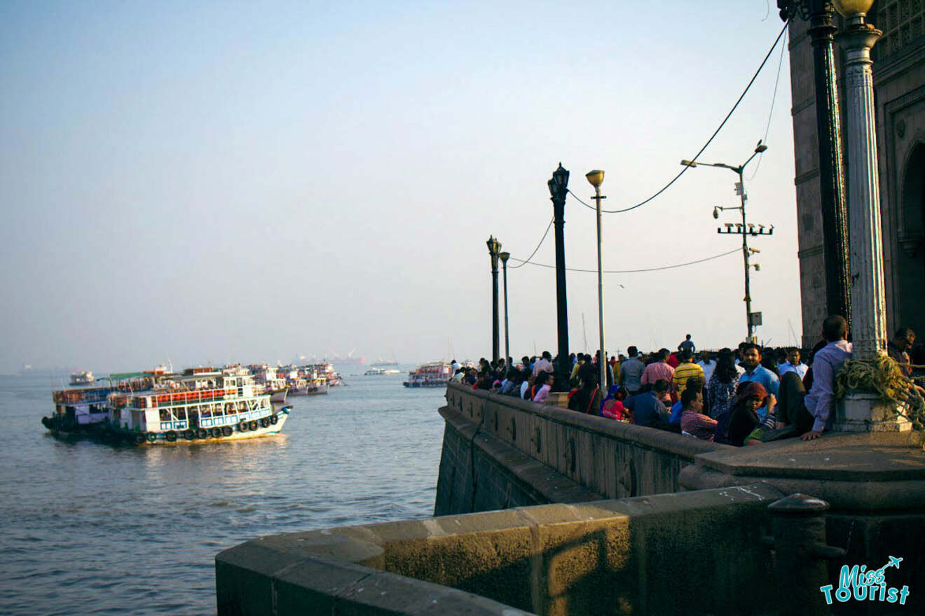 A crowd of people stand along a stone waterfront, watching multiple boats on the water under a clear sky.