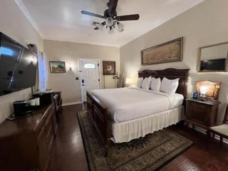 A bedroom with a large bed, two wooden nightstands, a wooden dresser with a TV on top, a ceiling fan, a wall mirror, and framed pictures. The room has wooden floors and a patterned rug under the bed.