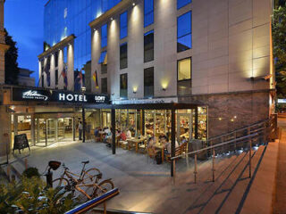 Exterior view of a hotel entrance at dusk with a glass-fronted restaurant and an outdoor seating area, bicycles parked nearby, and illuminated signs indicating the hotel name and restaurant.