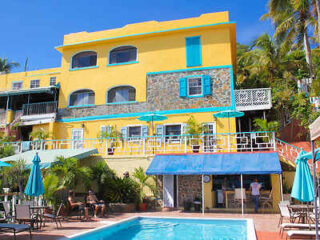 A vibrant yellow building with blue accents stands next to a pool with outdoor seating and surrounding palm trees.