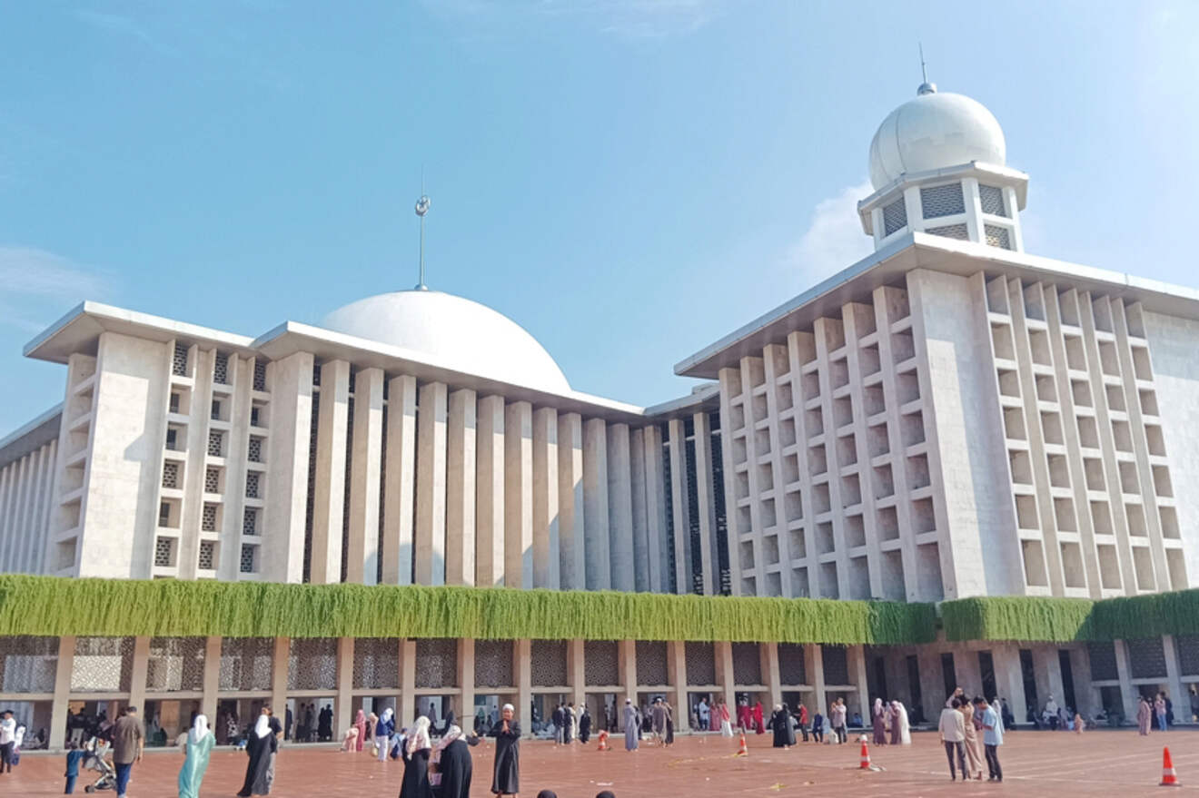 Large white mosque with a prominent domed roof and tall minaret. People are gathered around the entrance. The building features columns and lush green plants. Clear blue sky in the background.