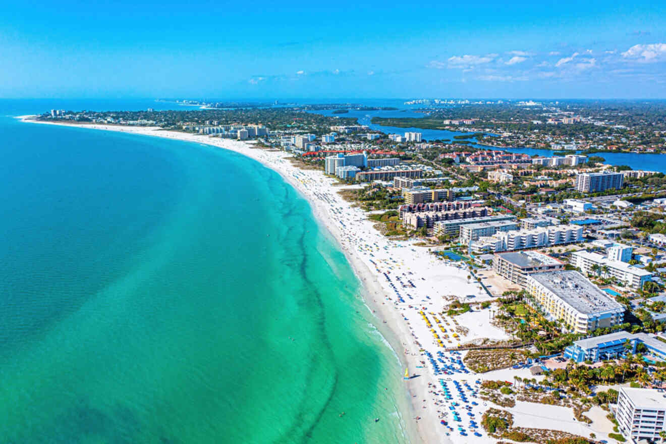 Aerial view of a pristine white sand beach with clear turquoise waters, lined with resort buildings and beachgoers, stretching far into the distance under a sunny sky.