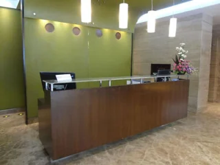 A wooden reception desk with two computers is situated in a modern office lobby. The background features a green wall, hanging lights, and a flower arrangement on the desk.