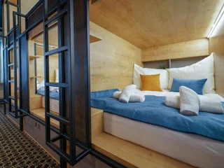 A cozy wooden room with a double bed dressed in blue and white linens, neatly rolled towels, and a yellow accent pillow. The bed area is sectioned off with black metal framework.