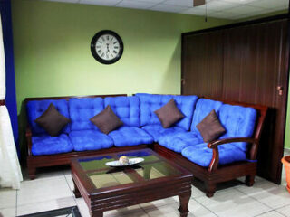 A living room with a blue cushioned corner sofa, three brown cushions, a wooden coffee table, a clock on the wall, and lime-green and blue walls.