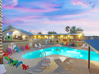 A backyard with a swimming pool surrounded by lounge chairs and colorful seating, with a sunset sky in the background, adjacent to a house.