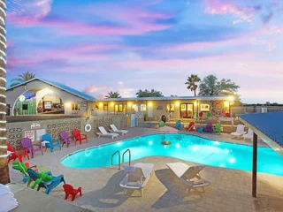 A backyard with a swimming pool surrounded by lounge chairs and colorful seating, with a sunset sky in the background, adjacent to a house.