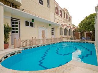 A serene outdoor swimming pool surrounded by a beige building with several windows and doors, featuring a geometric tile pattern at the bottom. A single white door is on the left wall.