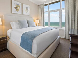 A bedroom with a large bed featuring white linens and a blue throw, flanked by two nightstands with lamps. Large window with sea view, two framed artworks above the bed. Neutral-toned decor.