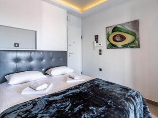 A modern bedroom with a double bed, white bedding, and a dark velvet blanket. There's a painting of an avocado on the wall and soft ceiling lighting.