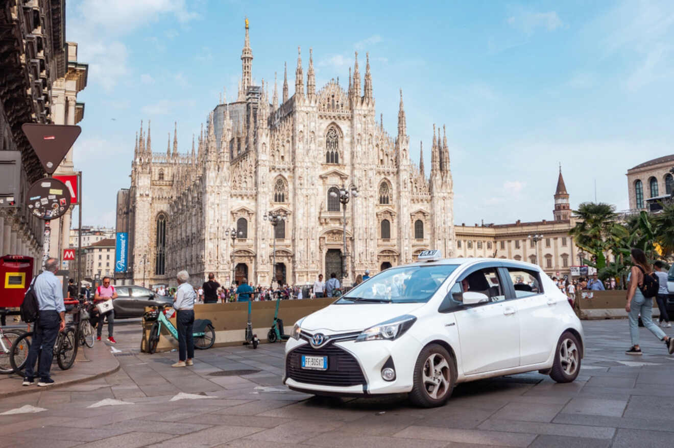 A white taxi is parked in front of an ornate, Gothic cathedral with people walking around and bicycles nearby.