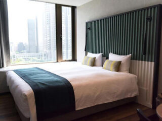 A modern hotel room features a large double bed with white linens and dark green accents, situated next to large windows offering a view of city buildings.