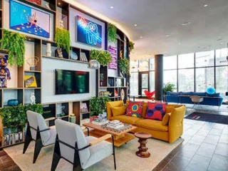 A modern living room with a yellow couch, two white chairs, a central coffee table, wall-mounted TV, and surrounded by plants and shelves with decor items.