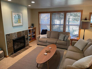 A cozy living room with two beige sofas, a wooden coffee table, a fireplace, a bookshelf, and large windows with blinds letting in natural light.