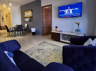 A modern living room with a blue sofa, a television mounted on a wall, and a dining area in the background. The room has contemporary decor with a large area rug and a painting on the wall.