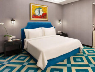 A neatly made bed with white bedding in a modern hotel room featuring blue and white geometric carpet, grey walls, a flower vase on a nightstand, and framed artwork above the headboard.