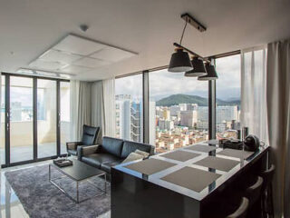 A modern apartment features a living area with a black leather sofa and a coffee table, a kitchen island with stools, and large windows offering a city skyline view.