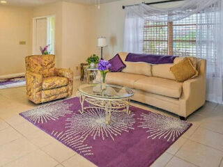 A beige sofa with purple and gold cushions stands beside a floral armchair in a living room with a purple rug and glass coffee table. A window with white sheer curtains is in the background.